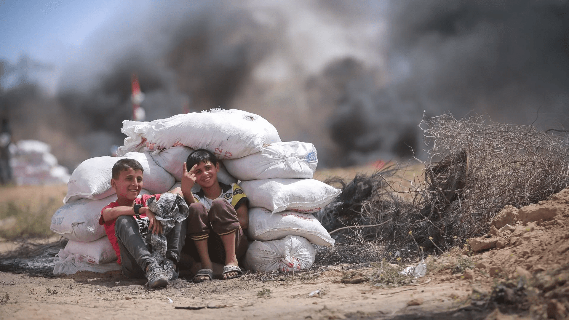 Two boys sit sheltering against a pile of sandbags as dark smoke rises behind them