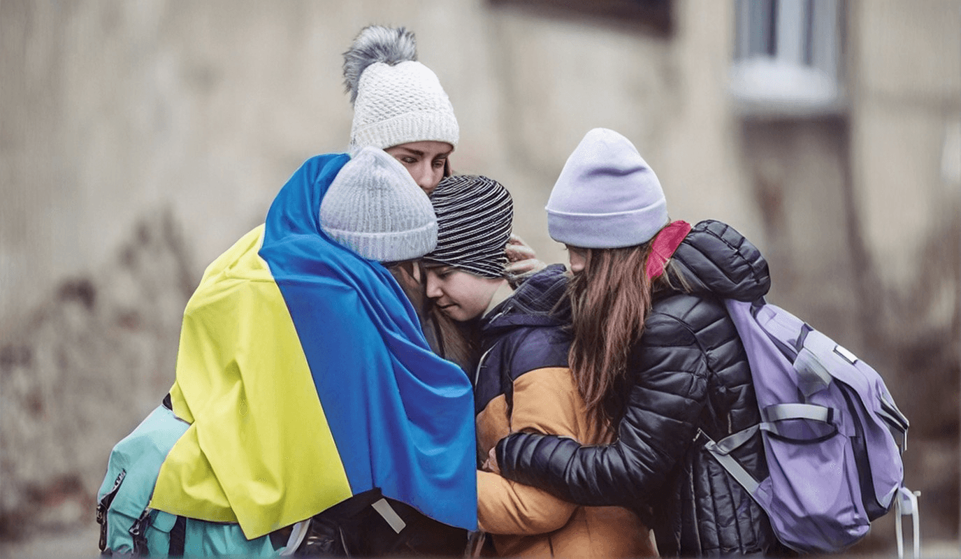 Four young women in winter hats huddle in a tight embrace, one wrapped in a Ukrainian flag
