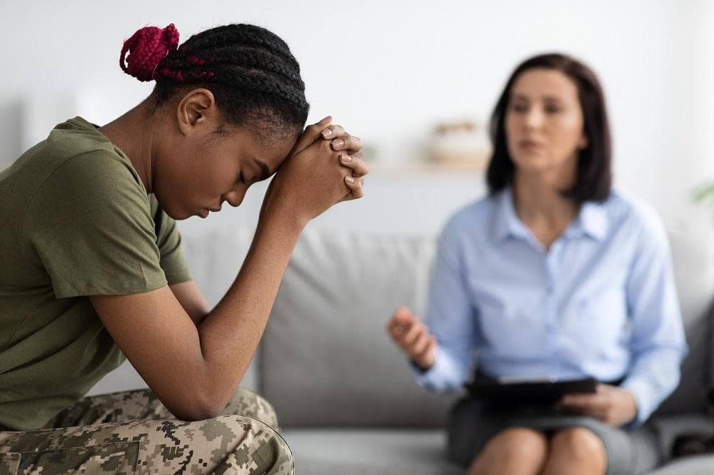 A young woman in military fatigues sits with her head bowed on her clasped hands while a therapist talks with her